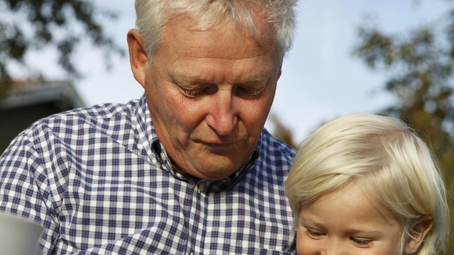 En bestefar og et barnebarn titter på et nettbrett sammen ute i fint vær.