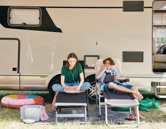 En jente og en gutt sitter i solstoler utenfor en campingbil og ser på hvert sitt nettbrett.