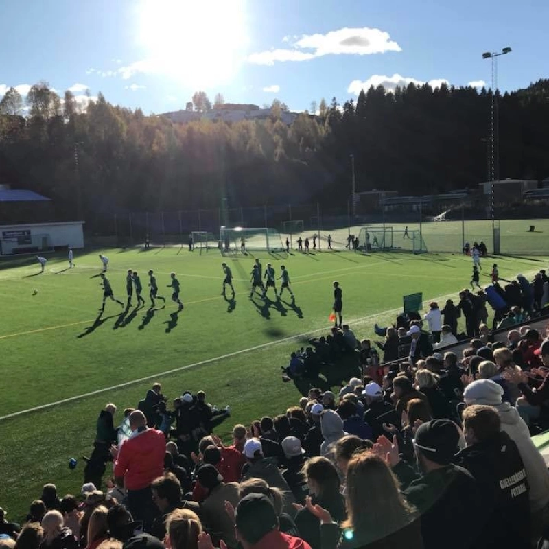 Bilde av en kamp som gjenomføres på en fotballbane med solskinn.